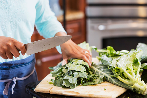 Cropped view of a mature African-American woman standing in the kitchen, cutting collard greens with a large knife.  The vegetable is common in southern cooking.