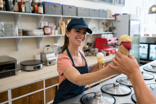 Female worker serving an ice cream to an unrecognizable customer at the ice cream parlor looking very happy and smiling