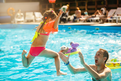 Happy active family young father and his cute daughter adorable toddler girl playing in a swimming pool jumping into the water enjoying summer vacation