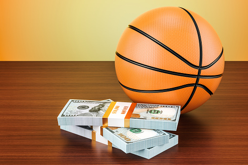 Sport bets. Basketball ball with dollar packs on the wooden table, 3D rendering