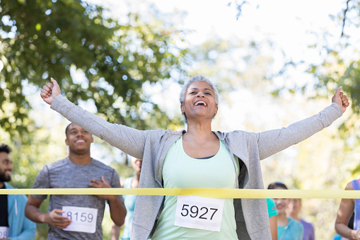 A smiling senior woman is running in a charity race.  She raises her arms before  breaking the ribbon and crossing the finish line first.
