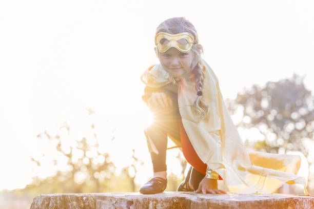 Little girl wearing a cape and mask is pretending to be a superhero. She's wearing a cape and mask, and is crouched down with the sun setting behind her as she plays outdoors.