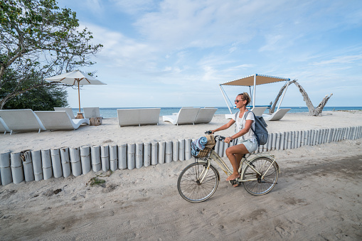 Cheerful young woman on vacations cycling by the beach in the morning, people travel holidays concept.