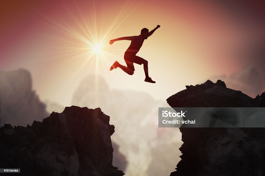 Silhouette of young man jumping over mountains and cliffs at sunset. Jumping Stock Photo