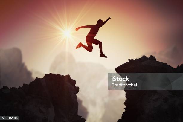 Silhouette Of Young Man Jumping Over Mountains And Cliffs At Sunset Stock Photo - Download Image Now