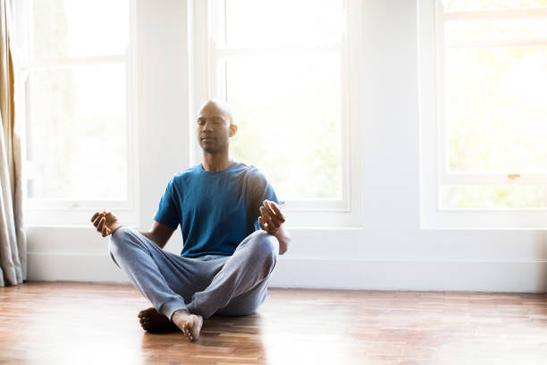 Man practicing yoga in lotus position at home Mid adult man practicing yoga in lotus position. Full length of male is exercising on hardwood floor. He is wearing sports clothing. cross legged stock pictures, royalty-free photos & images