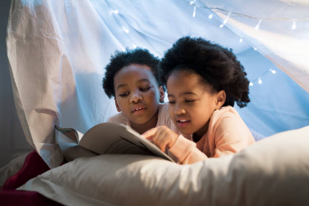 Little girls reading story book while lying on bed. Close-up of children are resting together at home. Sisters are relaxing in decorated tent.