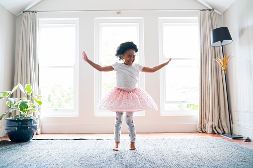 Full length of happy girl performing ballet dance at home. Female is wearing tutu while practicing. She is dancing against windows.