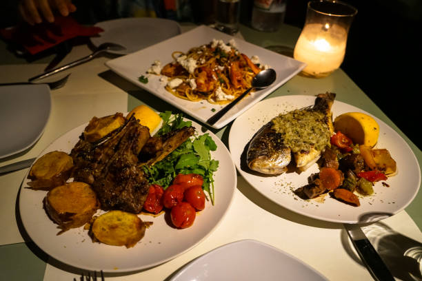 istock Delicious candle light dinner table serving fresh grilled lamb and whole sea bream fish with roasted potato, tomato, carrot, pepper, mushroom, zucchini, rocket salad and yellow lime; and seafood spaghetti pasta on white plate 915152578