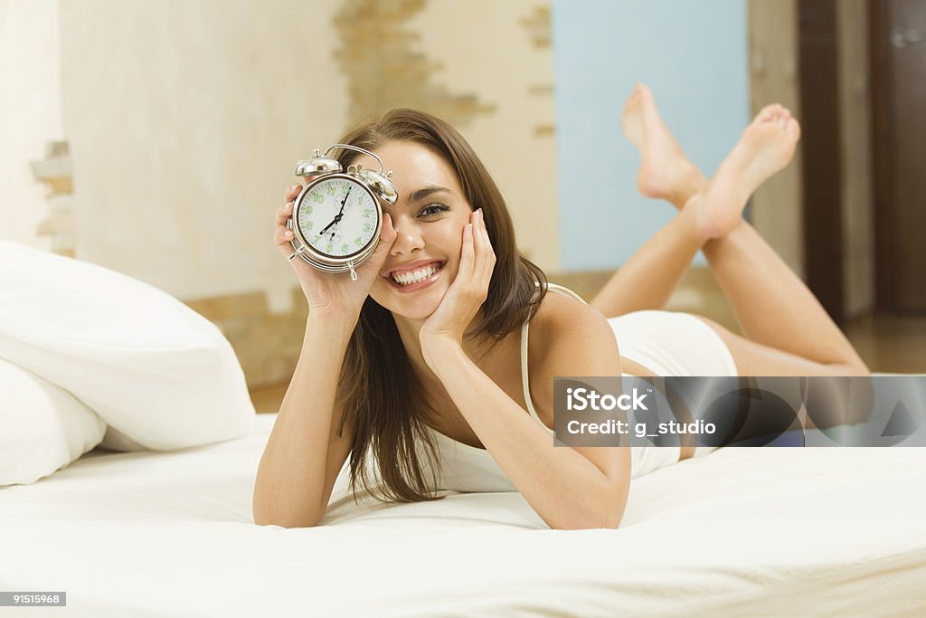 A young woman lying on a bed and holding up an alarm clock Young woman with alarmclock on the bed at morning Adult Stock Photo