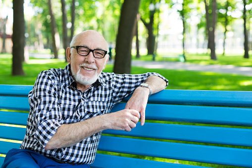 Smiling senior man outdoors. Elderly man in eyeglasses, wearing casual clothes, sitting on the bench in the park, posing on camera, copy space
