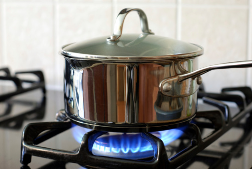 An old, covered, charred, blackened pot burning on a burner on an electric stove. The small flame is on the outside of the pot rising from the burner.