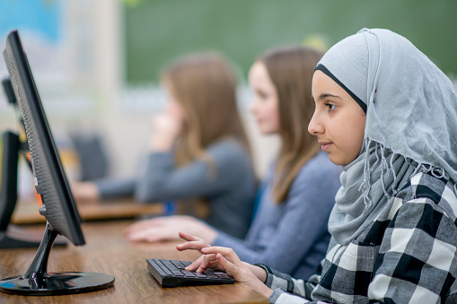A Muslim girl is indoors in her school's computer lab. She is wearing a head scarf. The girl is concentrating while typing on the computer keyboard.
