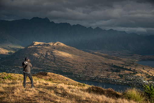 Young male photographer taking photo in photographing posture with mountain scenery during golden hour sunset in Queenstown, South Island, New Zealand. Travel and photography concept