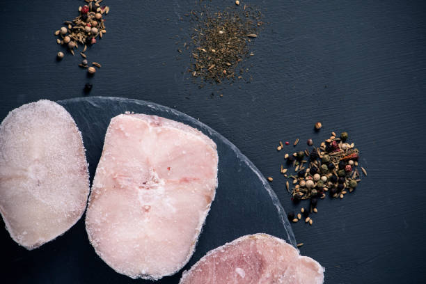 frozen hake cuts on a slate tray high angle view of some frozen hake cuts on a round slate tray, placed on a dark gray surface with some piles of different spices, such as different kinds of peppercorns hake stock pictures, royalty-free photos & images