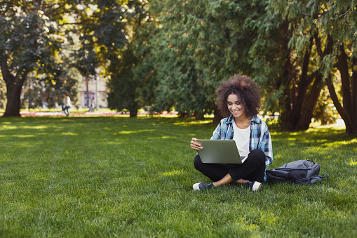 Joyful casual african-american student girl with laptop outdoors. Smiling woman sitting on grass with computer, surfing the net or preparing for exams. Technology, education and remote working concept