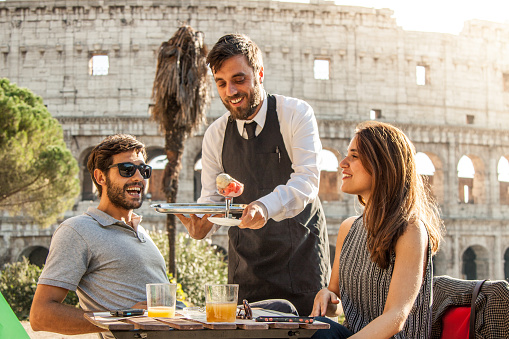 Elegant waiter serving a glass of colorful icecream on a try to a young happy couple in bar restaurant in front of colosseum in rome at sunset
