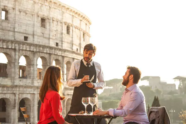 Photo of Elegant waiter writing orders on notebook serving a young happy couple in bar restaurant in front of colosseum in rome at sunset