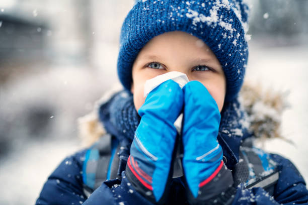 Portrait of little boy aged 8 going to school on winter day. The boy has runny nose and is blowing hos nose with a tissue.\n