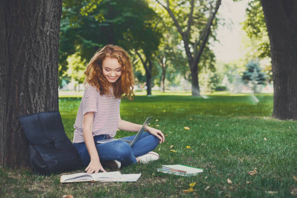 istock Redhead female student outdoors looking at book and using laptop sitting under a tree 915218128