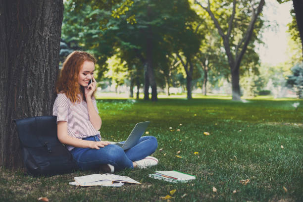 istock Redhead student girl in casual outdoors talking on mobile and using laptop sitting under a tree 915218172