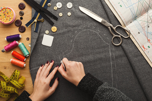 Sewing workshop or fashion designer at work. Top view on female hands marking cloth square with soap piece. Messy table with dressmaker accessories and patterns, copy space