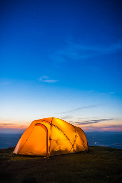 Stars shining above illuminated tent high on mountain after sunset Stars twinkling in a deep blue summer dusk sky over silhouetted mountain ridges and a bright orange illuminated dome tent. wales mountain mountain range hill stock pictures, royalty-free photos & images