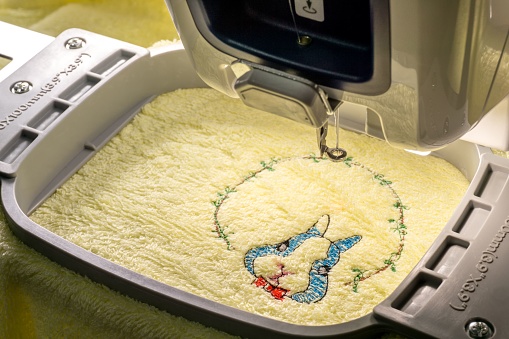 Workspace of embroidery machine embroidering blue cartoon  rabbit and wreath on yellow towel, copy space on the left, close up picture
