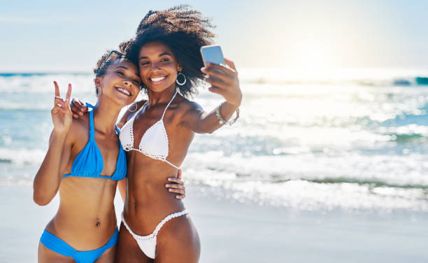 Stay beachy! Shot of two young women taking selfies together at the beach black women in bathing suits stock pictures, royalty-free photos & images
