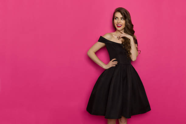 Elegant Young Woman Is Looking Away, Smiling And Thinking Beautiful young woman in elegant black cocktail dress is holding hand on chin, looking away and thinking. Three quarter length studio shot on pink background. cocktail dress stock pictures, royalty-free photos & images
