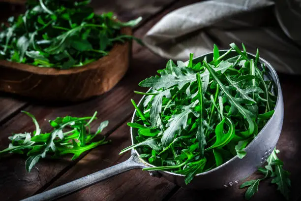 Fresh organic arugula in an old metal colander shot on rustic wooden table. This vegetable is considered a healthy salad ingredient. Predominant colors are green and brown. Low key DSRL studio photo taken with Canon EOS 5D Mk II and Canon EF 100mm f/2.8L Macro IS USM