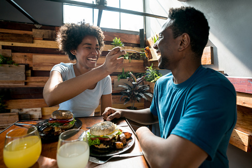 Happy african american couple at a restaurant and woman feeding her boyfriend both laughing and smiling