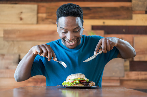 Portrait of a black happy man starving and eating a delicious hamburger using a fork and knife