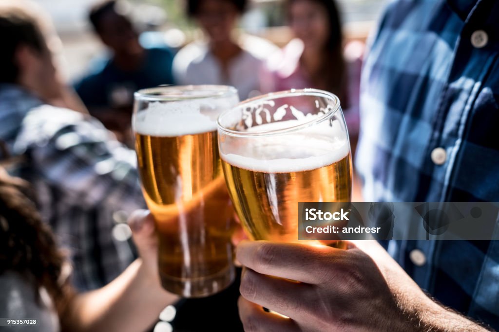 Close up of a customers at a bar holding a beer and making a toast Close up of a customers at a bar holding a beer and making a toast with people having fun at the background Beer - Alcohol Stock Photo