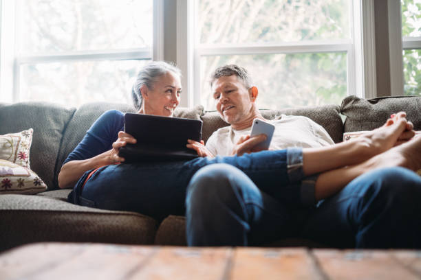 Mature Couple Relaxing with Tablet and Smartphone A couple in their 50's relax in their home on the living room couch, enjoying reading and surfing the internet on their mobile touchscreen phones and computer tablet. comfortable stock pictures, royalty-free photos & images