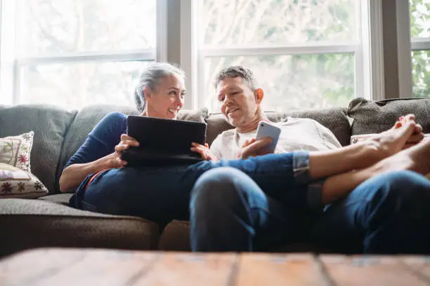 A couple in their 50's relax in their home on the living room couch, enjoying reading and surfing the internet on their mobile touchscreen phones and computer tablet.