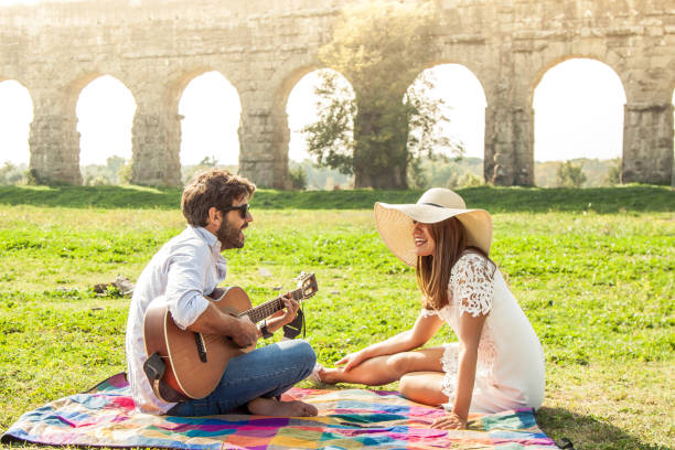 bella ragazza con cappello con un giovane che suona la chitarra nel parco in una giornata di sole. coppia felice seduta su una colorata coperta da picnic di fronte alle rovine dell'acquedotto romano a roma - ponte di strumento musicale foto e immagini stock