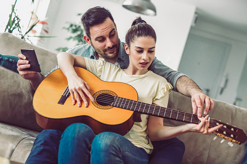 Young couple playing guitar on couch indoor. Woman is learning to play a guitar.