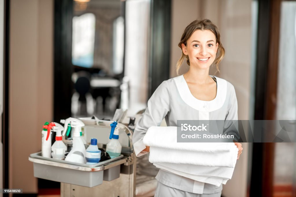 Chambermaid in the hotel corridor Portrait of a young woman chambermaid holding a towel standing with maid cart full of cleaning stuff in the hotel corridor Hotel Stock Photo