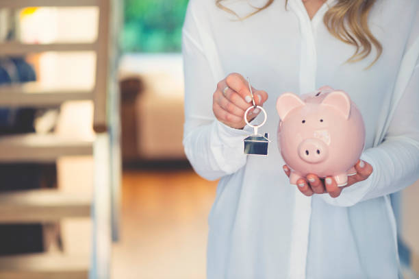 Woman holding house keys and a piggy bank Woman holding house keys and a piggy bank. The key ring is house shaped. There is a home interior in the background. Home ownership and savings concept. Copy space. australia house home interior housing development stock pictures, royalty-free photos & images