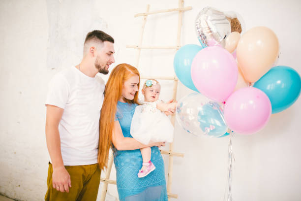 a young family of three people, mom's dad and daughter's one year old stands inside the room. holding a balloon in her hand, a woman with red long hair is holding a child in her arms dressed in a white dress. birthday party of a child - long hair red hair women men imagens e fotografias de stock