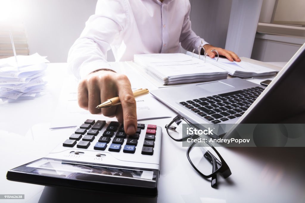 Businessman Calculating Tax Using Calculator Midsection Of Businessman Calculating Tax Using Calculator With Laptop On Desk In Office Accountancy Stock Photo