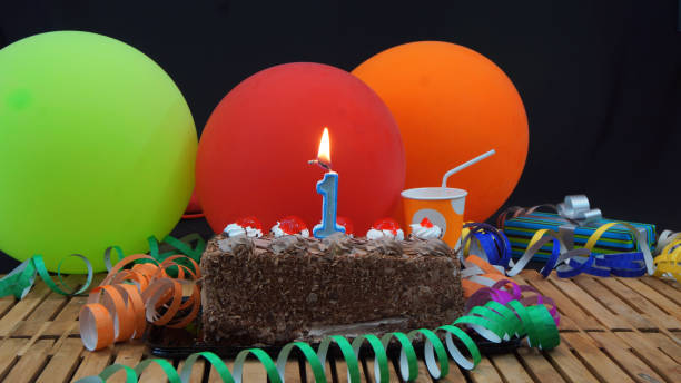 istock Chocolate 1 birthday cake with candles burning on rustic wooden table with background of colorful balloons, gifts, plastic cups and streamers with black background 915524176