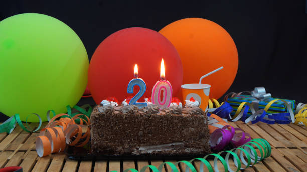 istock Chocolate 20 birthday cake with candles burning on rustic wooden table with background of colorful balloons, gifts, plastic cups and streamers with black background 915527716