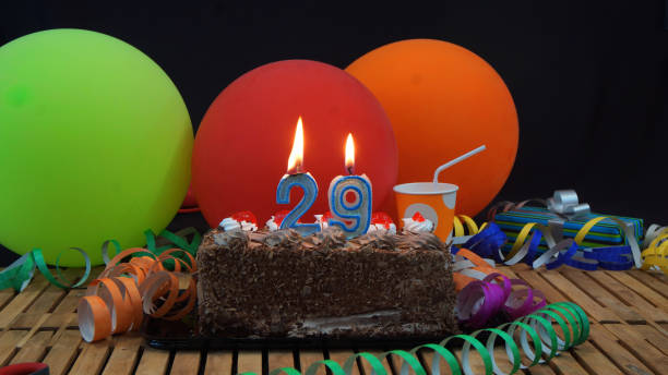 istock Chocolate 29 birthday cake with candles burning on rustic wooden table with background of colorful balloons, gifts, plastic cups and streamers with black background 915528094