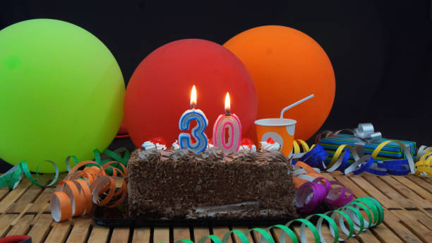 istock Chocolate 30 birthday cake with candles burning on rustic wooden table with background of colorful balloons, gifts, plastic cups and streamers with black background 915528276