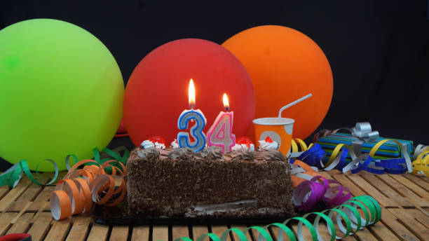 istock Chocolate 34 birthday cake with candles burning on rustic wooden table with background of colorful balloons, gifts, plastic cups and streamers with black background 915528340