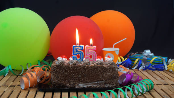 istock Chocolate 55 birthday cake with candles burning on rustic wooden table with background of colorful balloons, gifts, plastic cups and streamers with black background 915534228