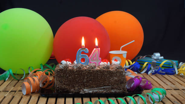 istock Chocolate 64 birthday cake with candles burning on rustic wooden table with background of colorful balloons, gifts, plastic cups and streamers with black background 915534756