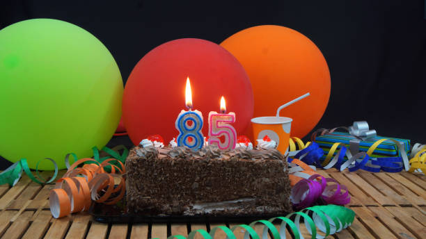 istock Chocolate 85  birthday cake with candles burning on rustic wooden table with background of colorful balloons, gifts, plastic cups and streamers with black background 915538128